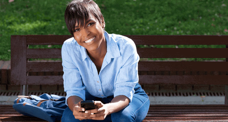 A smiling woman sitting on a bench holding her cellphone with both of her hands