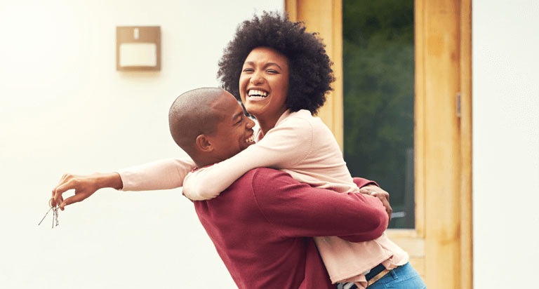 A smiling man lifts and hugs a smiling woman in front of a door while the woman holds keys in her right hand