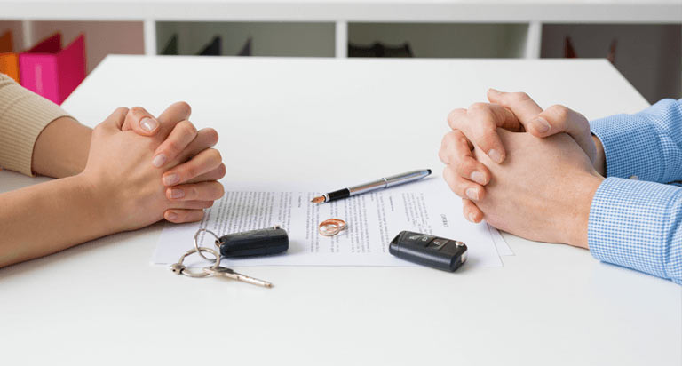 Two people sit at a table with their hands clasped and on the table are two car key FOBs, two rings, a pen, and a sheet of paper