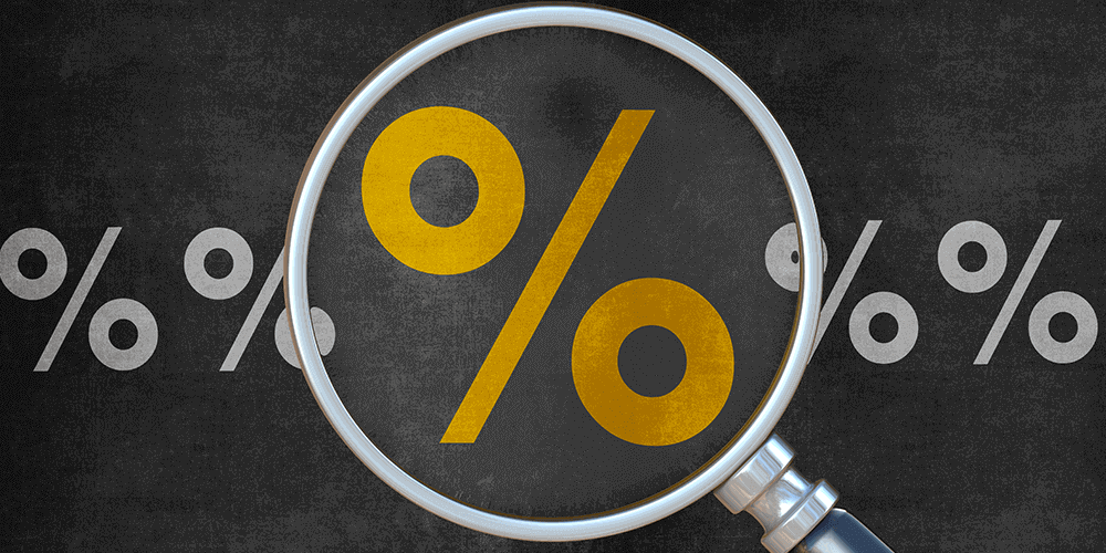There is a row of grey percentage rate symbols on a chalkboard background. In the center, a magnifying glass is making one yellow percentage rate symbol look larger than the rest.