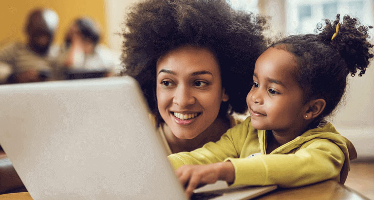 A woman and a child smile at a laptop screen while the child types on the keyboard of the laptop