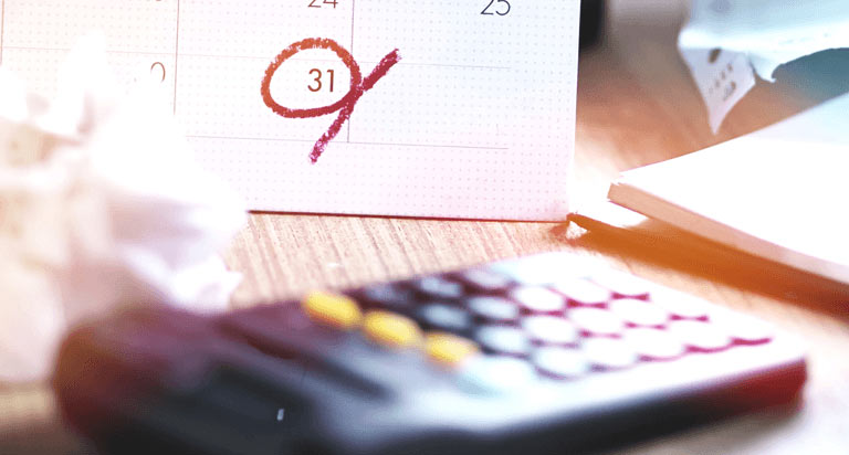 A calendar with the 31st circled in red with a notebook, a calculator, and a crumpled ball of paper sit in the foreground