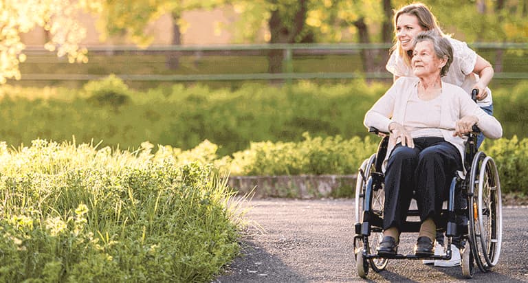 A smiling young woman pushing a smiling older woman in a wheelchair through a garden
