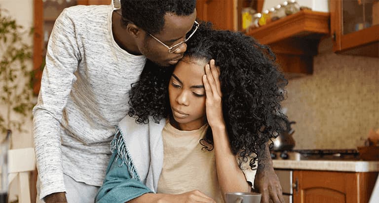 A man in glasses holds a woman who is holding her head with her left hand and seems overwhelmed in the kitchen