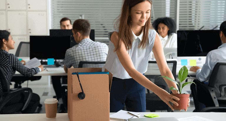 A woman sets up her new desk at work. She puts a small plant on her desk, next to her computer. Several coworkers sit behind her.