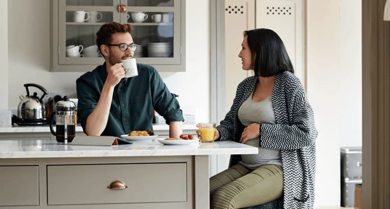 A man wearing glasses and holding a mug and a pregnant woman look at each other while having breakfast