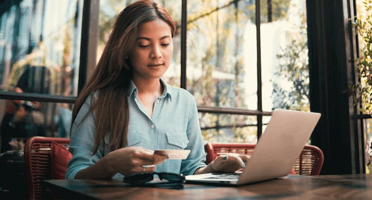 A woman sitting at a table inside a shop is reading her credit card information with a laptop in front of her.
