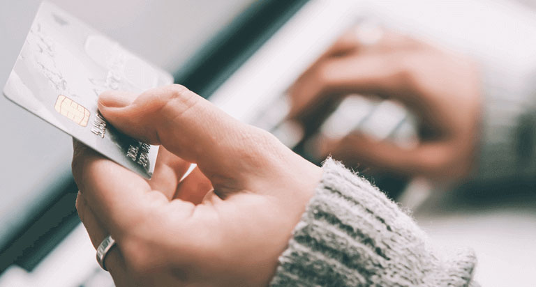 A person is holding a credit card in their hand while typing on a laptop.
