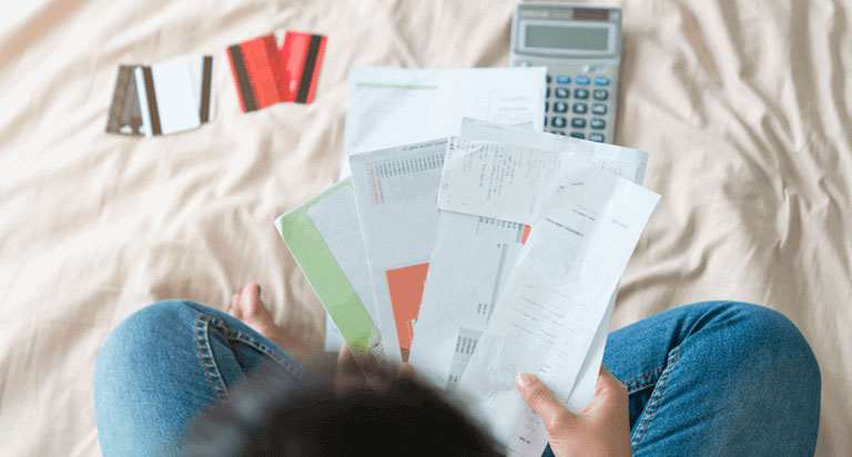 A calculator and 5 credit cards lay spread out on a bed. A person holds up a handful of financial papers above.