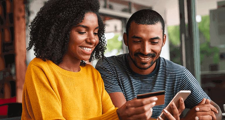 A woman holding a credit card in her right hand and a cellphone in her left hand while smiling and sitting next to a man who is also smiling