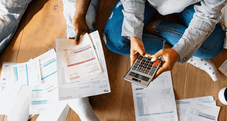 A woman holding and typing on a calculator with both hands sitting next to a man holding an invoice with his left hand