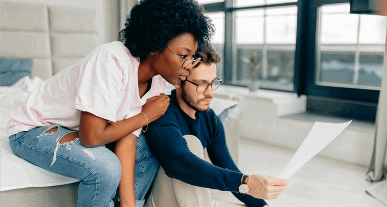 A couple looks over a financial paper together. The man is seated, while the woman leans on him looking over his shoulder.