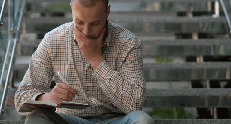 A man focusing while writing in a notebook on his lap