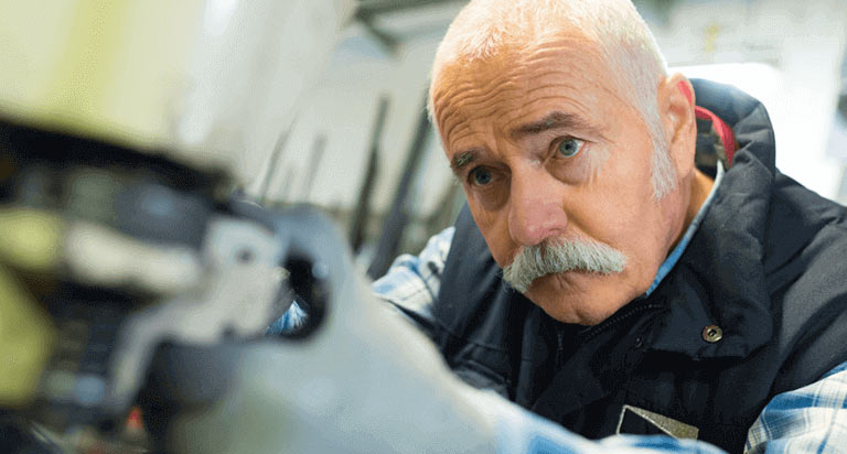 A man in a workshop works on a piece of machinery. He is wearing a striped button down and a work vest.