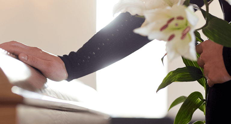 A person putting their right hand on a closed casket while the left hand holds a flower