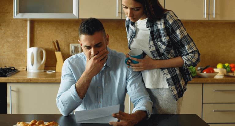 A man sits at a kitchen table looking at a paper and looks distressed. A woman stands over him also looking at the paper.