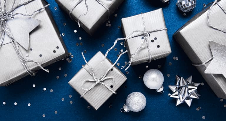 Silver boxes wrapped with silver twine and silver stars sitting on a blue background with silver ornaments, silver bows and glitter around them