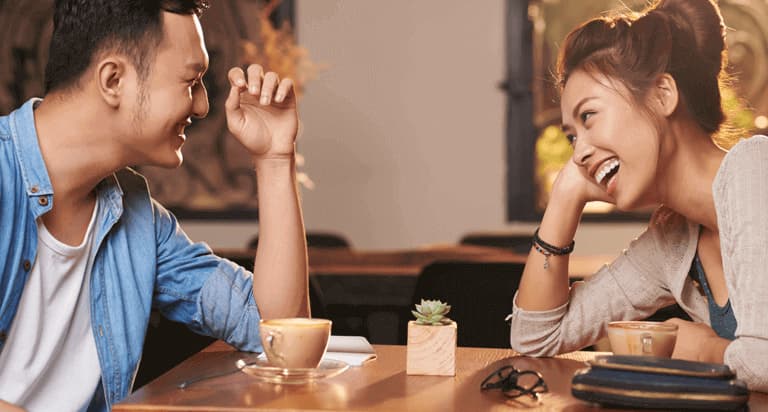 A man and a woman looking at each other and smiling while drinking coffee