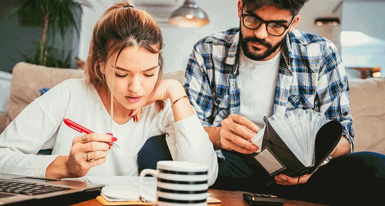 A woman and a man both look down at papers on a table while the woman holds a red pen in her right hand and the man holds an accordion folder