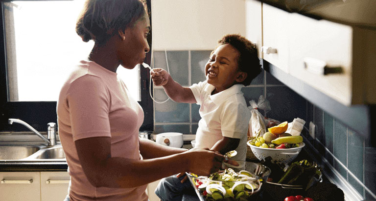 A mom and her child smile at each other in the kitchen. The child sits on top of the counter, while feeding his mom with a spoon.