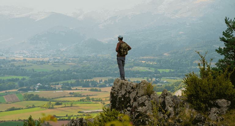 A man standing on the top of a cliff on a hiking trail looking down at a city