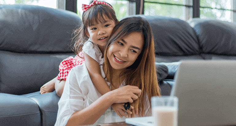 A mom smiling while using her laptop; her daughter is smiling while she hugs her mom.