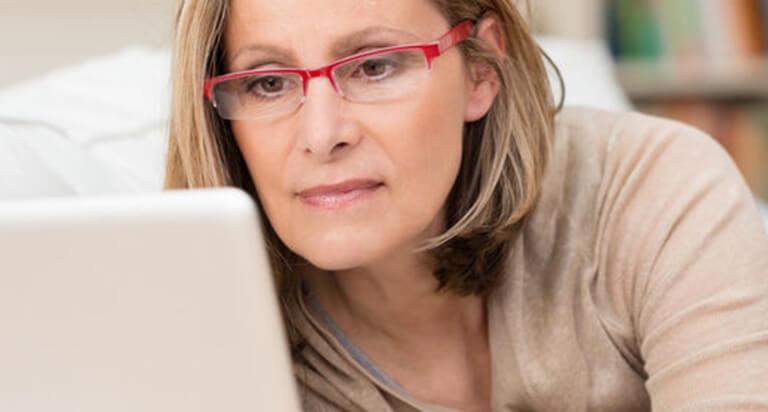 A woman wearing red glasses and a beige shirt is reading something on her laptop.