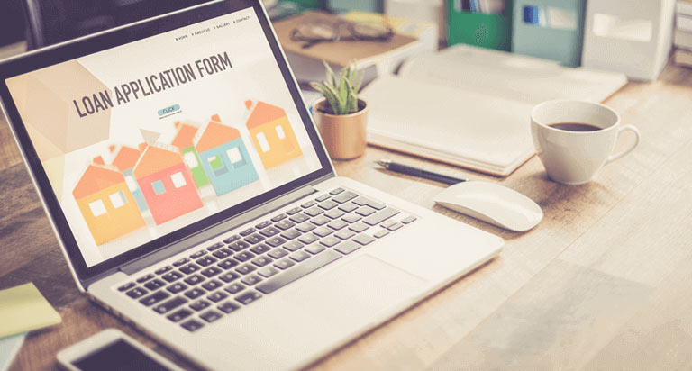 An opened laptop showing a loan application page on the screen with a cellphone, sticky notes, a succulent, a notebook, a pen, and a white coffee mug on the table beside the laptop