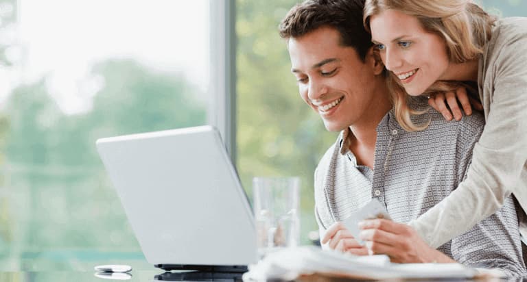 A smiling woman leaning on a man who is smiling while looking at his laptop and holding a credit card with his left hand