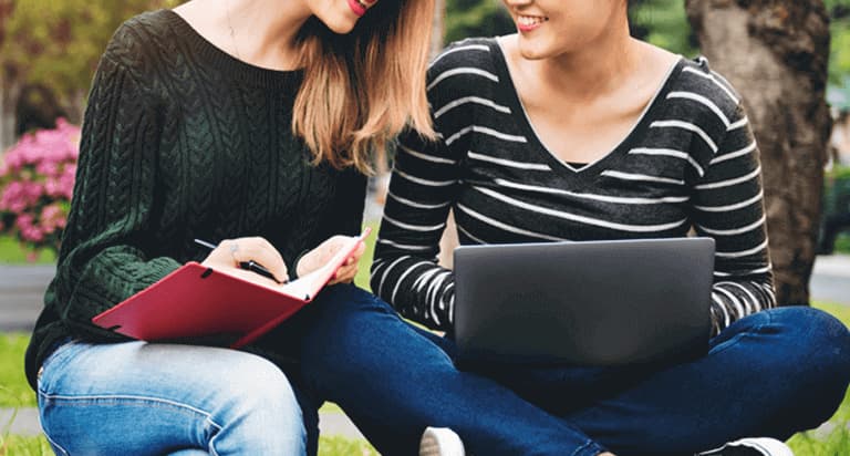 Two women sitting in the grass while the woman on the left holds a book and the woman on the right holds a laptop