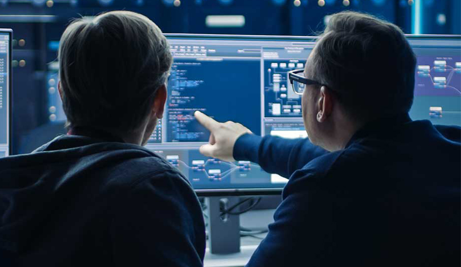 Two IT professionals seen from behind, looking at a computer monitor displaying code and network diagrams in a high-tech server room.