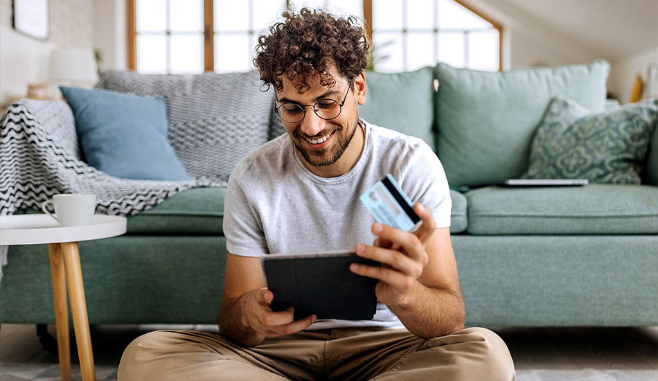 Jovem faz compras no seu tablet na sala de estar enquanto segura um cartão de crédito na mão.