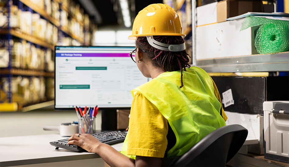 Mujer con casco de seguridad revisa el computador dentro de una gran bodega de paquetería