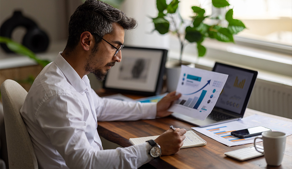 Hombre escribe notas en su cuaderno mientras observa documento con gráficos en su escritorio