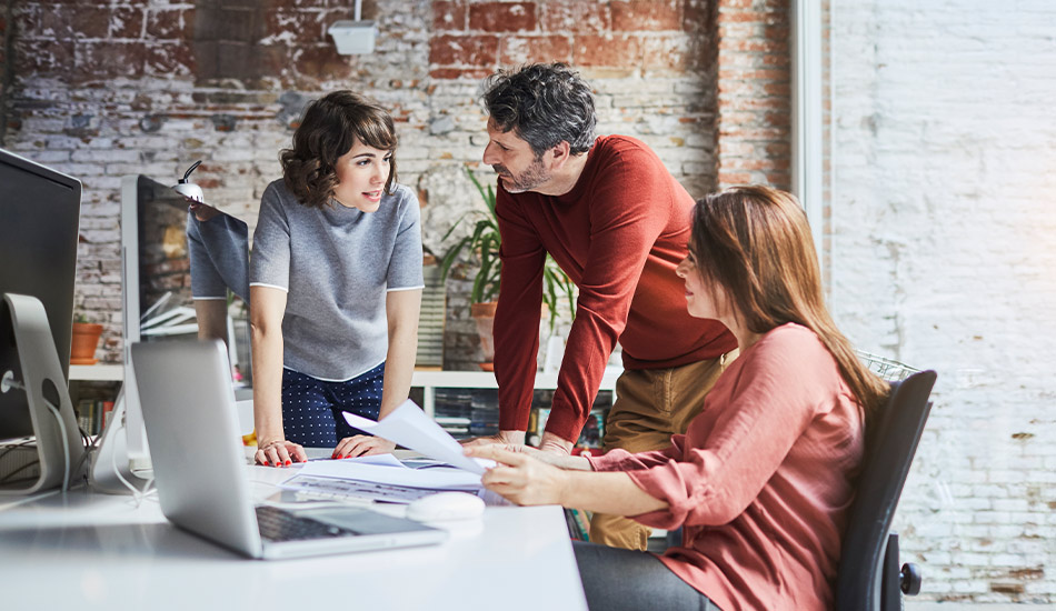 Tres empleados conversan informalmente en una oficina mientras revisan documentos