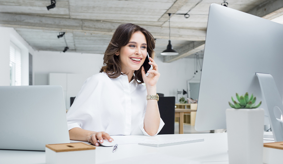 Mujer en amplia oficina realiza llamada telefónica en su escritorio mientras también revisa información en su computador