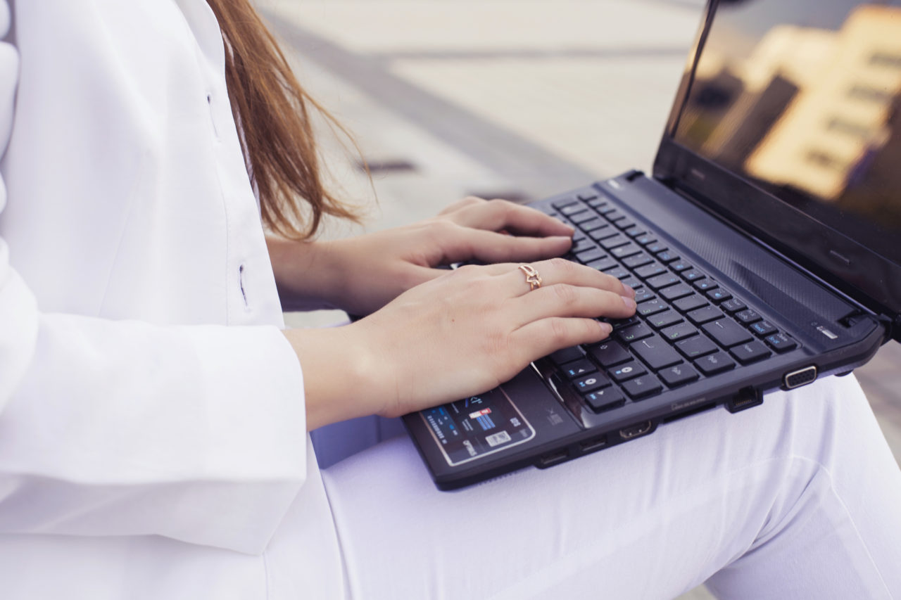 Close-up of woman sitting outside in all white with laptop on her lap, hands touching the keyboard