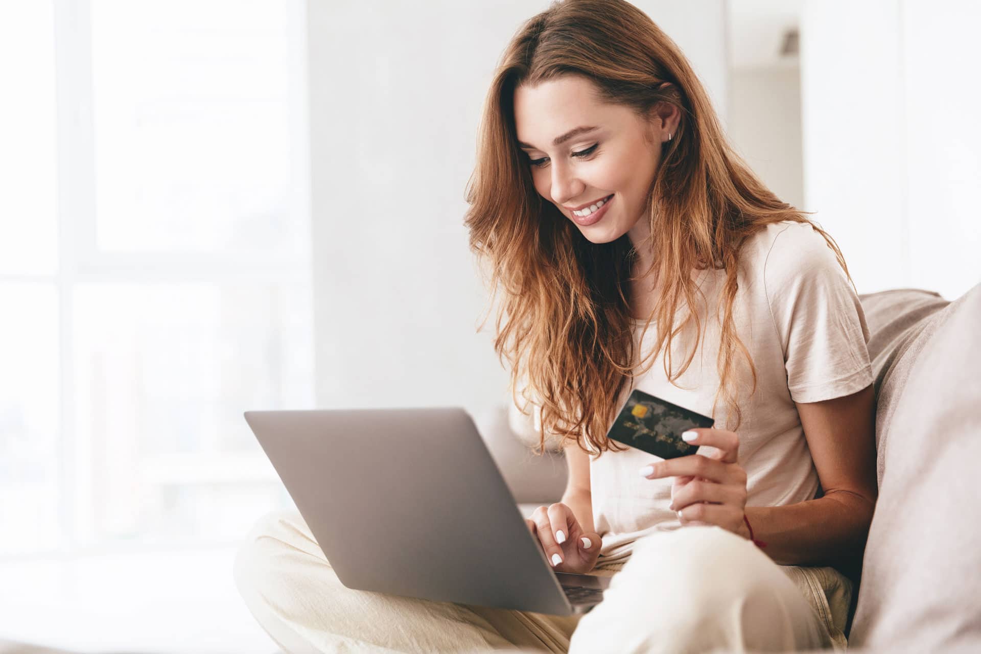 Woman with long brown hair in white outfit smiling, sitting on couch with laptop on lap and credit card in hand.