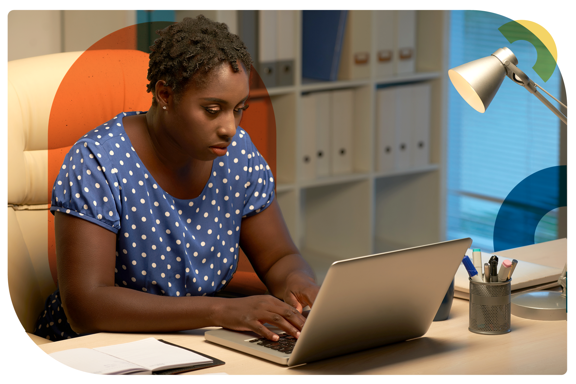 An african american woman in a blue polka dot blouse is sitting at a desk in front of a laptop typing and focused in on the screen