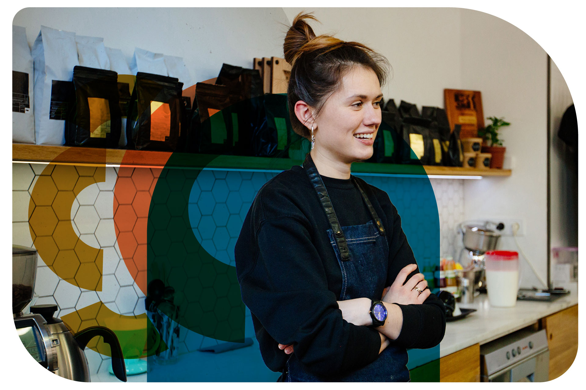 A woman with her hair tied up with silver drop earrings and wearing an apron is standing up against the counter of what appears to be a coffee shop. The shelf behind her has coffee bags lining the entire shelf. She is looking off camera and smiling with her arms crossed.