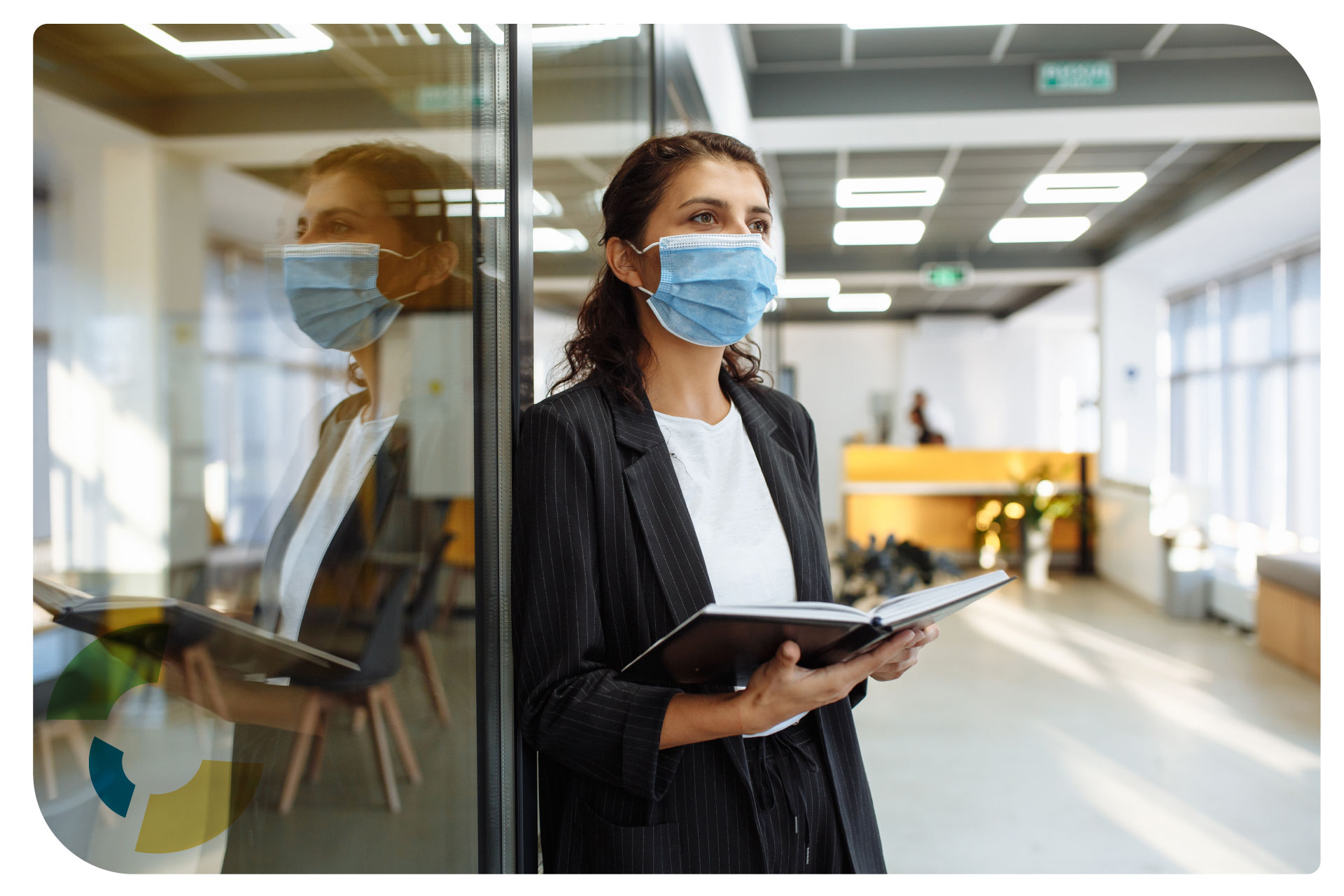 Woman with mask on standing in front of a conference room with a notebook ready to take notes